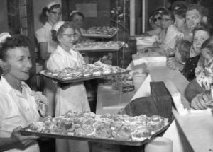 Four bakers are holding up large trays laden with fresh cream puffs at a State Fair midway concession stand, while an eager crowd stands waiting in anticipation on the other side of the counter.