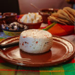 A small wheel of white cheese with tortillas and salsa in the background