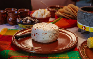 A small wheel of white cheese with tortillas and salsa in the background