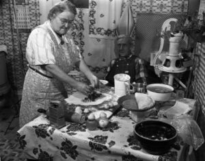Woman wearing an apron fills a pasty in her Iowa County, Wisconsin kitchen. A man wearing coveralls is seated at the table watching. (Courtesy of Wisconsin Historical Society)