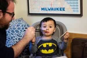 3-year old Axel Neilsen sits in a high chair being fed by his father.