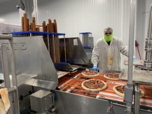 Bernatello’s employee Nelson Aguilar watches over a machine that slices long pepperoni sticks. The pieces fall onto the crusts below. (Photo by Jana Rose Schleis)