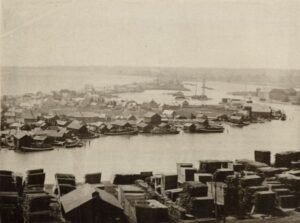 Elevated view of fishing village on Jones Island, Milwaukee in 1892. Several fishing boats are docked along the shoreline. Large piles of lumber are in the foreground across from the island.