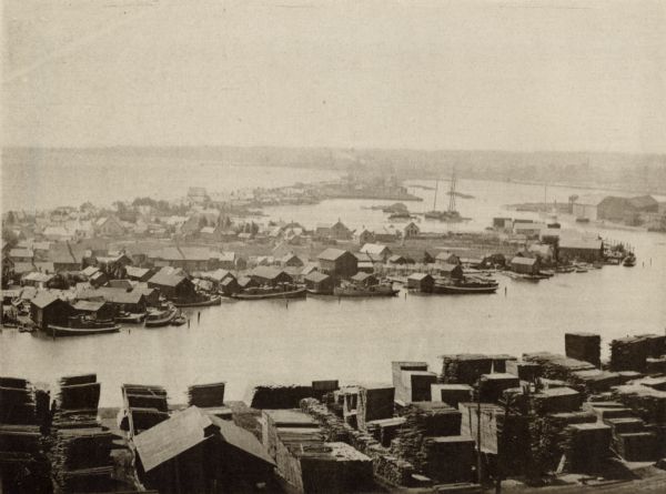 Elevated view of fishing village on Jones Island, Milwaukee in 1892. Several fishing boats are docked along the shoreline. Large piles of lumber are in the foreground across from the island.
