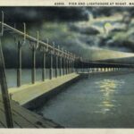 Hand-colored postcard view of a pier and lighthouse at night. The moon shines through clouds, and light shines in two directions from the beacon on top of the lighthouse. A ship is on the horizon. Caption reads: "Pier and Lighthouse at Night, Manitowoc, Wis." (Courtesy of Wisconsin Historical Society)