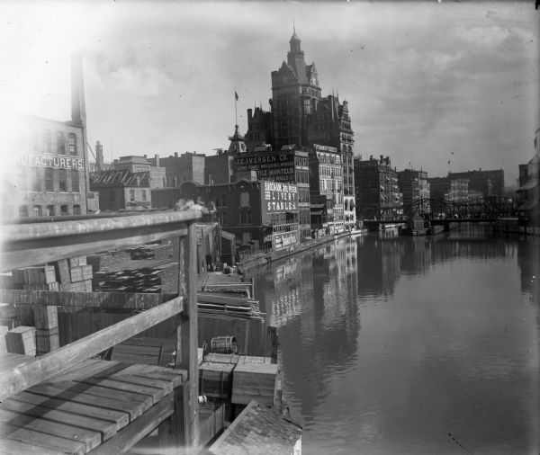 View of downtown Milwaukee, Wisconsin around 1898 from pier or dock of buildings along the Milwaukee River, including a building with a clock tower. Signage on brick exteriors reads "J.C. Iverson Co. / Picture Frames, Mouldings, Mirrors," "B. Mock & Son Hotel Pfister Livery Stables," "The Worlds Breakfast, Quaker Oats," and "Espenhain Dry Goods Co. Wholesale & Retail, Carpets & Draperies." (Courtesy of Wisconsin Historical Society)