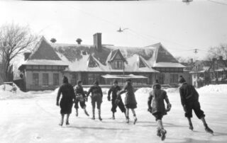 Seven children are ice skating toward the rear of a long, one-story building with half timber details under the eaves and a massive central chimney in Milwaukee, Wisconsin in 1928. Piles of snow line the large rink. Snow has been cleared up to the building, which appears to be a commercial building, or possibly a clubhouse. Electric lights are hanging from wires suspended over the rink. There are houses along a street in the background on the right. (Courtesy of Wisconsin Historical Society)