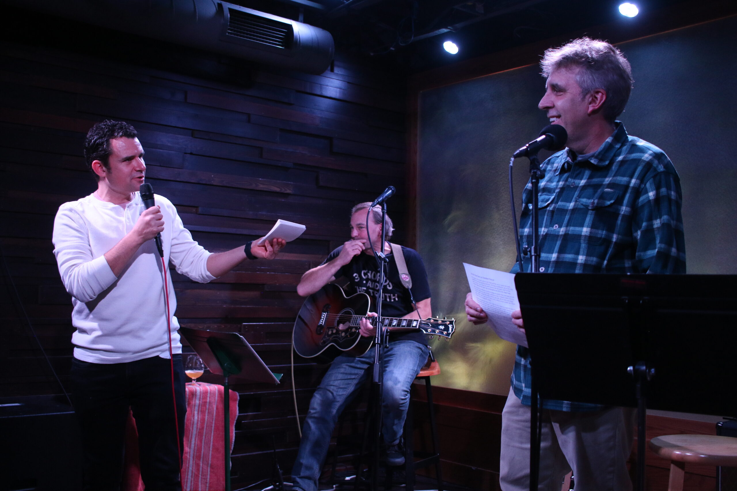 Author B.J. Hollars, left, talks with writer Ken Szymanski after he read his essay “As the Sun Sets on Summer Soccer” while musician Derrick Black is seated with a guitar. Szymanski shared his story on stage at The Lakely on Nov. 14, 2024. Hollars co-hosted the event put on by WPR’s “Wisconsin Life” and the Chippewa Valley Writers Guild. <i>Rich Kremer/WPR</i>