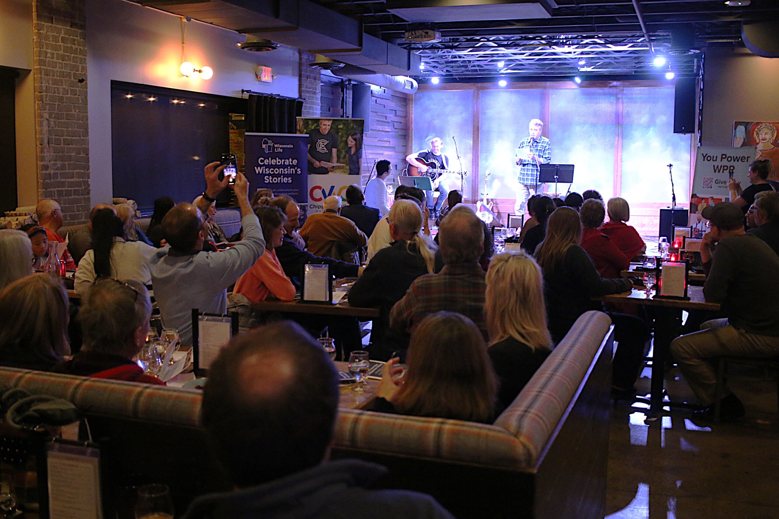 Musician Derick Black, left, and writer Ken Szymanski, right, of Eau Claire, on stage at The Lakely in Eau Claire on Nov. 14, 2024. They performed to a packed house for a storytelling event hosted by WPR’s “Wisconsin Life” and the Chippewa Valley Writers Guild. <i>Rich Kremer/WPR</i>