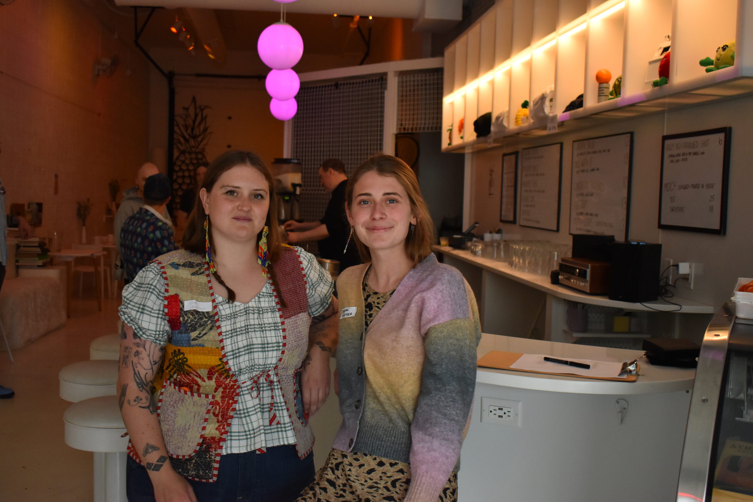 Two women stand together inside a modern café with a bar counter, illuminated by purple hanging lights and menu boards on the wall behind them.