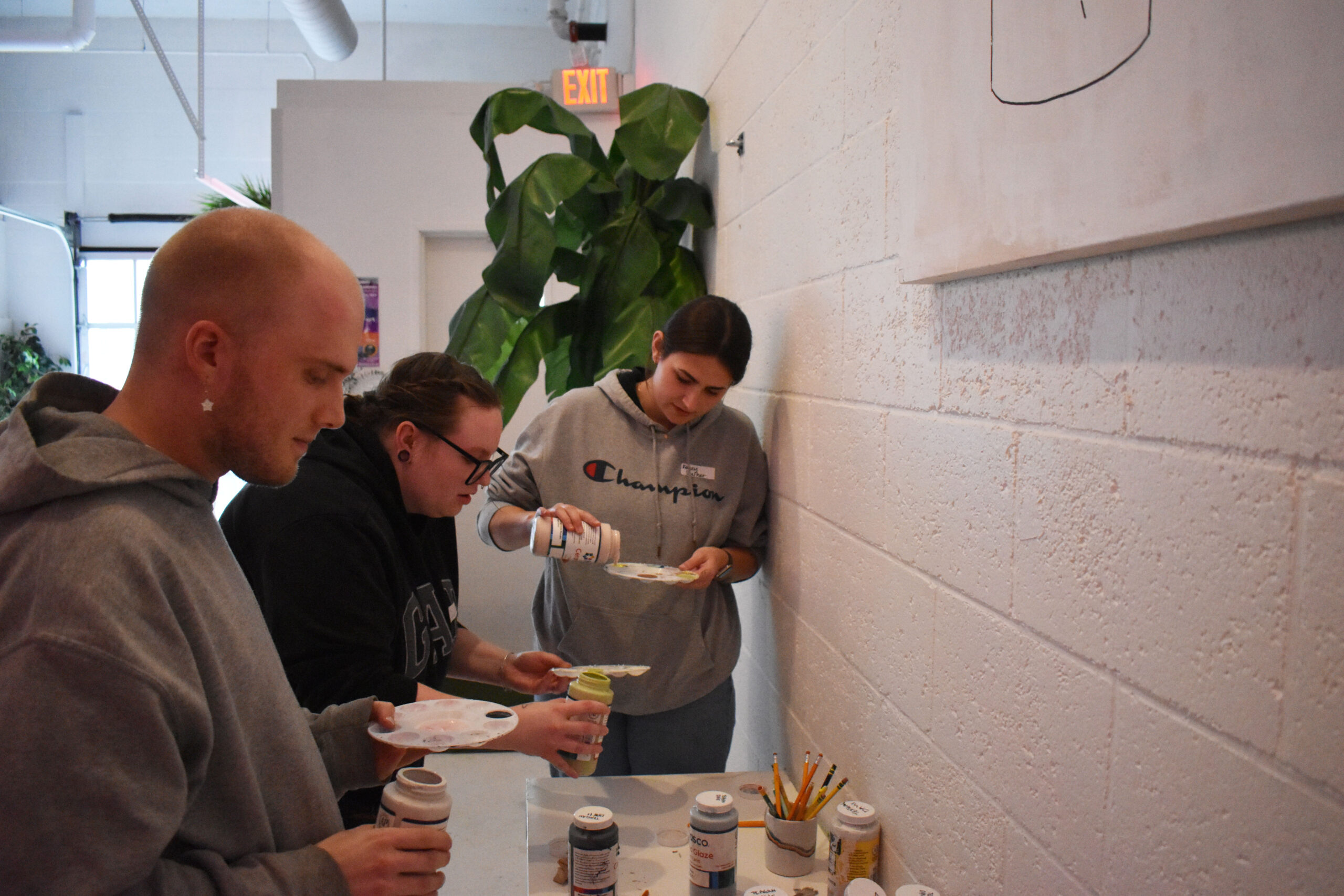 Three people stand at a table with paint bottles and palettes, preparing to paint on canvases in a well-lit room with white walls and a large plant.