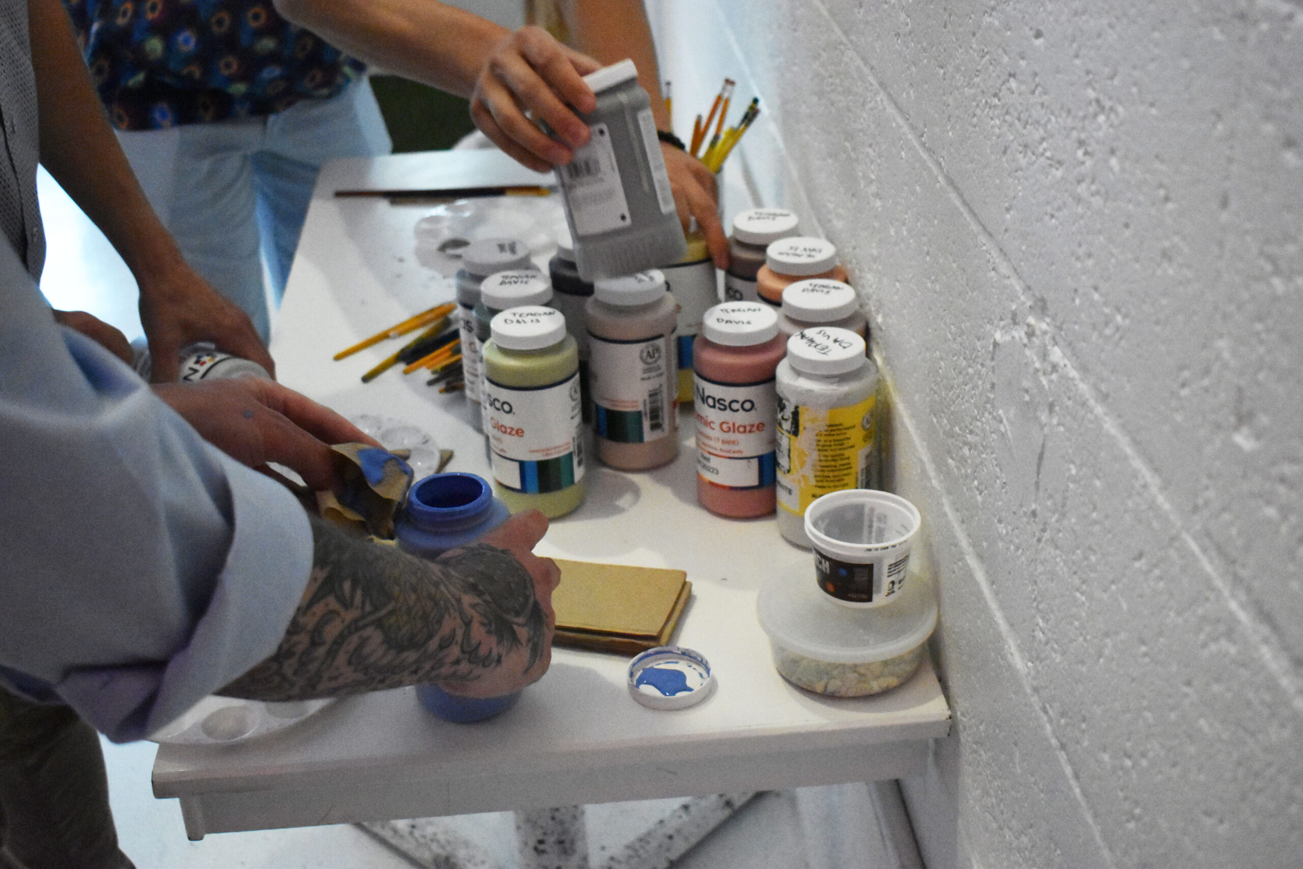 People gather around a small table with jars of ceramic glaze, paintbrushes, pencils, and art supplies, preparing materials for an art project.